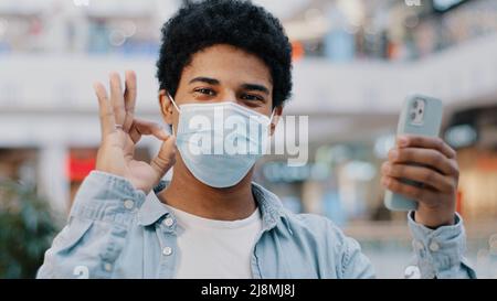 Ritratto africano dominicano ragazzo malato uomo in maschera medica protettiva con telefono che mostra ok gesto alla fotocamera raccomanda buona app medica felice con onli Foto Stock