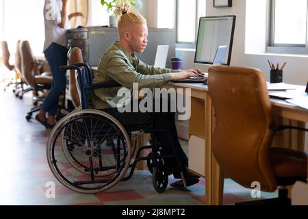 Albino afroamericano uomo d'affari medio adulto seduto su sedia a rotelle mentre si usa il computer portatile alla scrivania Foto Stock