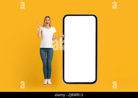 Felice scioccata giovane europea donna con bocca aperta in bianco t-shirt punti dito su enorme telefono con schermo vuoto Foto Stock