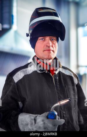 Il saldatore maschio in abiti da lavoro con maschera sulla testa guarda direttamente nella macchina fotografica. Ritratto autentico di lavoratore in sala di produzione. Persone reali. Foto Stock