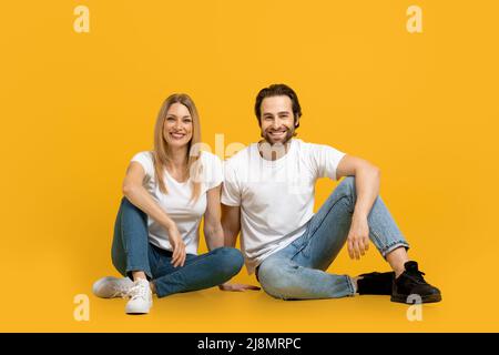Felice sicuro bel ragazzo millenario europeo con barba e donna in t-shorts bianco siedono sul pavimento insieme Foto Stock