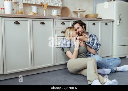 Sorridendo felice uomo europeo millenario con la moglie stoppia e bionda godere di momento tenero, abbraccio, seduto sul pavimento Foto Stock