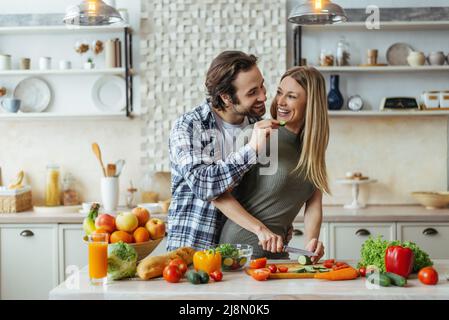 Felice marito millenario caucasico con stubble nutre moglie bionda, coppia hanno divertimento, signora preparare insalata Foto Stock