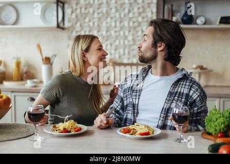 Felice felice bella coppia millenaria caucasica mangiare pasta insieme, divertirsi in cucina leggera interno, profilo Foto Stock