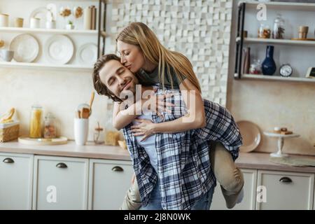 Soddisfatto ragazzo millenario europeo con stoppia tiene la donna bionda sulla sua schiena, signora bacia guancia, godere del tempo libero Foto Stock