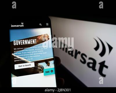 Persona che tiene lo smartphone con la pagina web della società satellite Inmarsat Global Limited sullo schermo di fronte al logo. Concentrarsi sul centro del display del telefono. Foto Stock