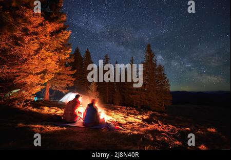 Campeggio notturno vicino alla foresta. Coppia di turisti che trascorrono una serata romantica all'aperto in falò sul prato. Sullo sfondo tende, pini, cielo stellato. Attività ricreative nella natura. Sfocatura del movimento. Foto Stock