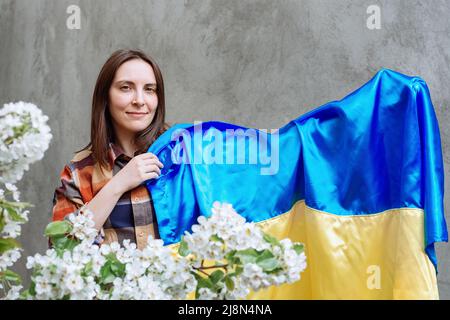 Bella ragazza tiene in mano la bandiera blu-gialla di stato dell'Ucraina su uno sfondo di cemento grigio città muro. Donna che indossa una camicia a quadri. Apple Blossoms sfocato in primo piano. Spazio di copia. Foto Stock