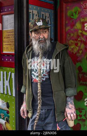 Un uomo pesantemente trafitto con una lunga barba intrecciata grigia, vestita casualy, si trova in una porta con una tazza di caffè nella sua h Foto Stock