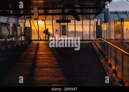 Tramonto alla stazione della ferrovia leggera Poplar Docklands nella zona est di Londra, un ciclista per la consegna di cibo lancia una silhouette. Foto Stock