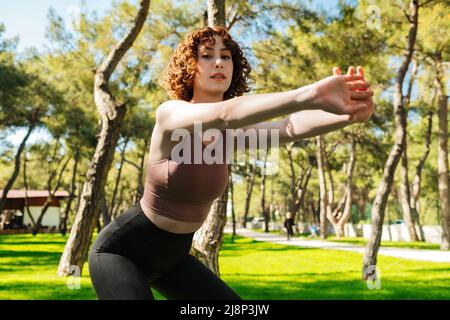 Una giovane donna rossa che dura nel tempo si allena al parco cittadino, all'aperto facendo squat profondi con le braccia distese. Donna sportiva che guarda la fotocamera. Sport c Foto Stock