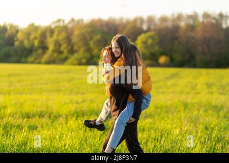 Madre giocosa che dà la figlia piggy giro indietro al campo verde. Entrambi ridendo e sembrano felici. Primavera in background foresta. Primo piano. Foto Stock