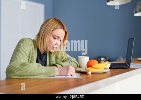 La giovane donna prende appunti al tavolo della cucina. Foto Stock