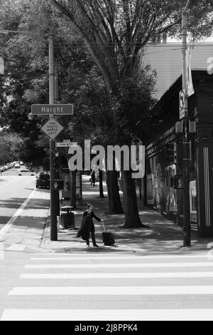 Giovane donna in attesa di attraversare la strada all'angolo di Masonic Avenue e Haight Street; Haight-Ashbury District a San Francisco, California, valigia. Foto Stock