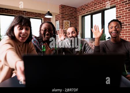 Persone multirazziali che sventolano la mano alla videoconferenza online su un portatile mentre si siede a casa. Diversi gruppi di amici che fanno un gesto di saluto sullo schermo del computer durante una conversazione su Internet. Foto Stock