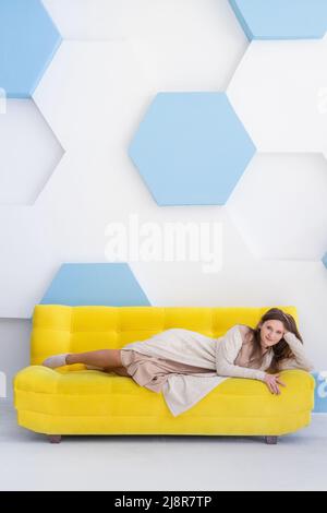 Giovane felice donna caucasica si trova su un divano giallo in interni luminosi con spazio copia, verticale. Foto Stock