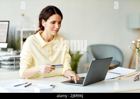Scatto orizzontale medio della giovane donna caucasica adulta seduta alla scrivania in ufficio utilizzando il portatile e la carta di credito per ordinare la consegna del cibo Foto Stock