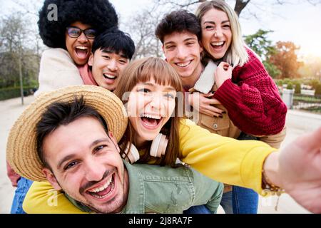 Gruppo allegro di amici felici che prendono selfie sorridenti in piggyback. Tre coppie che si divertono insieme all'aperto nel parco della città. Persone che godono Foto Stock