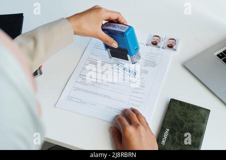 Vista ad alto angolo di una donna musulmana irriconoscibile che indossa hijab che lavora come ufficiale consolare seduto alla scrivania mettendo timbro approvato su carta documenti Foto Stock