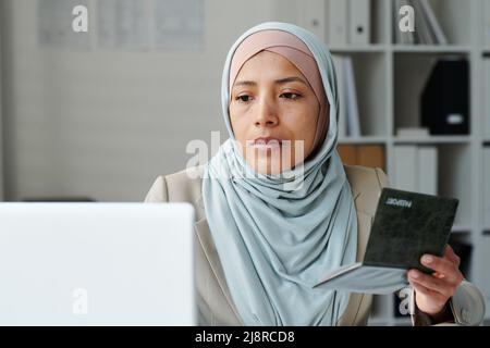 Ritratto di giovane donna musulmana seria adulto che indossa hijab seduta in ufficio ambasciata utilizzando un computer portatile per lavorare con i documenti Foto Stock