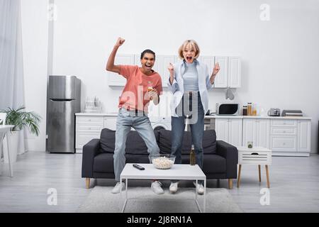 felice coppia multietnica saltare mentre si guarda lo sport partita in salotto Foto Stock