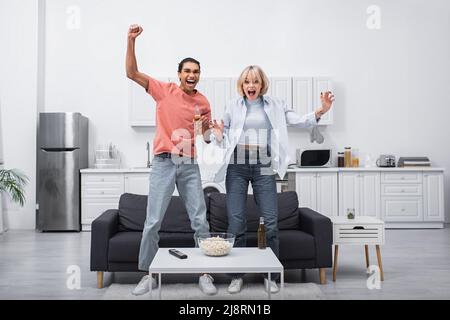 coppia multietnica eccitata saltando mentre si guarda lo sport partita in salotto Foto Stock
