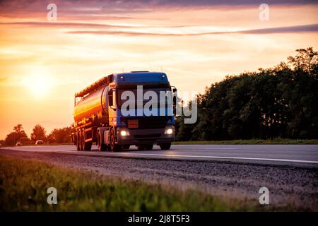 Un moderno camion blu con una petroliera semi-rimorchio trasporta merci pericolose sullo sfondo del tramonto serale. Il concetto di trasporto di Foto Stock