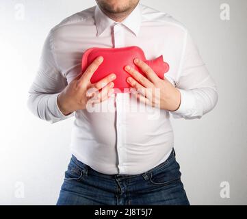 Un uomo in una camicia bianca tiene un cuscinetto riscaldante rosso con acqua calda sul petto. Trattamento dell'infiammazione dei polmoni e della bronchite mediante riscaldamento. Termostato Foto Stock