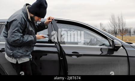 Maschio ladro burglar passando da sciocchi attraverso la crepa nella finestra di parcheggio auto vuoto, si apre la porta sbloccato, tooks smartphone e fugge scena con stol Foto Stock