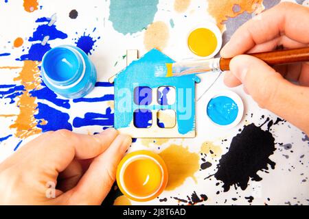 Vista dall'alto di mani pennello pittura un modello di legno piatto di una casa su un colorato sfondo di tessuto. Concetto di hobby e ricreazione. Foto Stock