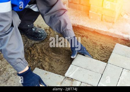 Il muratore in abiti da lavoro si siede sul marciapiede e stende le lastre di pavimentazione. Vista dell'uomo in attività all'aria aperta. Costruttore professionista fa organizzazione del territorio giorno d'estate. Scena reale. Foto Stock