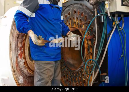 L'ingegnere in casco ispeziona e ripara le apparecchiature a gas del locale caldaie. Pulizia e manutenzione di caldaie industriali a vapore. Foto Stock