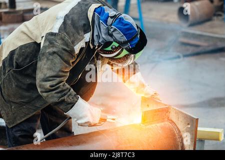 Saldatrice professionale in uniforme protettiva e maschere in sala di produzione. Flusso di lavoro autentico. La saldatrice lavora con metallo e scintille volare. Foto Stock