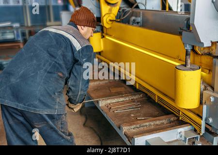 L'operaio taglia il metallo sulla macchina meccanica della ghigliottina nel locale di produzione. Attrezzature industriali per il taglio di metalli. Scena reale. Flusso di lavoro reale. Uomo al lavoro. Foto Stock