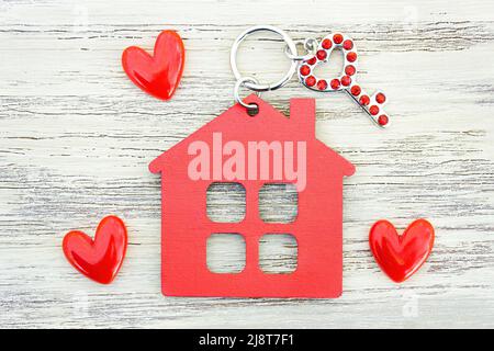 Piccola chiave a forma di cuore decorata con rhinestones rossi, portachiavi a forma di casa piatta e piccoli cuori rossi su uno sfondo di legno. Dolce casa romantica con Foto Stock