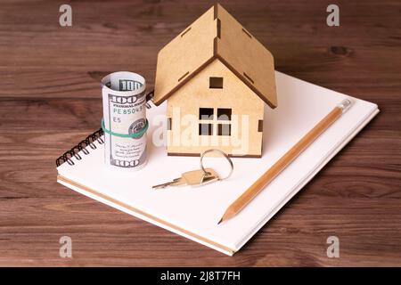 Piccola casa di legno compensato, rotolo di bollette del dollaro, chiave e matita su un taccuino di carta. Investire nel settore immobiliare. Foto Stock