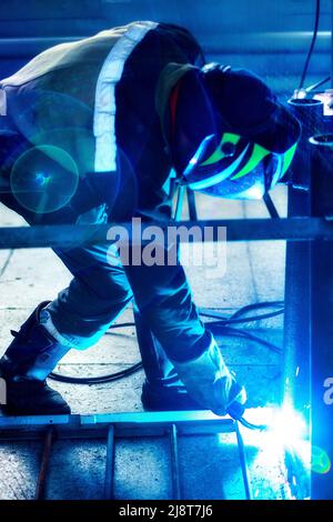 Saldatrice professionale in uniforme protettiva e maschere in sala di produzione. Flusso di lavoro autentico. La saldatrice lavora con metallo e scintille volare. Foto Stock