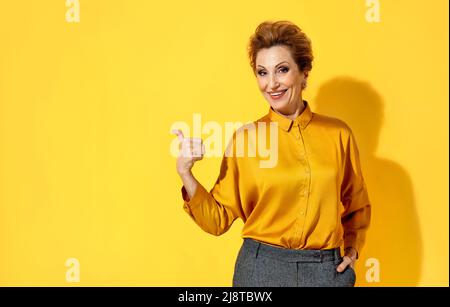 La donna sorridente punta a parte con il pollice, mostra sullo spazio di copia per il testo. Foto di attraente donna anziana in camicia gialla su sfondo giallo Foto Stock