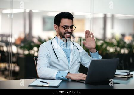 Felice bel giovane medico islamico maschio con barba in occhiali, cappotto bianco guarda il computer portatile, mano ondeggiante Foto Stock