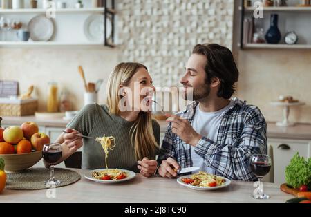 Sorridendo maschio caucasico giovane con stubble nutre la donna bionda, coppia hanno divertimento insieme in cucina interna chiara Foto Stock
