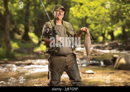 Sorridente pescatore maturo che tiene un pesce carpa e in piedi in un fiume nel bosco Foto Stock