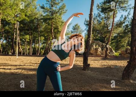 Donna rossa sportiva che indossa un reggiseno sportivo verde e pantaloni yoga blu che si esercitano prima di fare jogging al parco, allungando i muscoli all'aperto, sentendo la salute Foto Stock