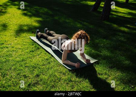 Giovane donna rossa attraente che indossa abiti sportivi che fanno esercizi in plancia sul tappeto yoga al parco cittadino, all'aperto. Concetto di vita sana e sport all'aperto Foto Stock