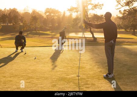 Giovane africano americano che tiene bandiera mentre gioca a golf con l'amico caucasico al campo da golf Foto Stock