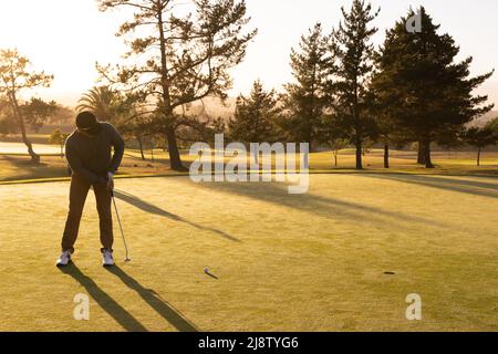 Tutta la lunghezza del giovane caucasico che colpisce la sfera di golf con il randello contro gli alberi e cielo chiaro al tramonto Foto Stock