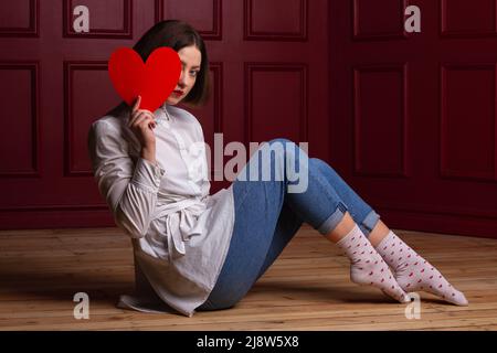 Donna dai capelli corti seduta lato alla macchina fotografica su pavimento di legno e sfondo rosso che tiene la forma del cuore rosso davanti a lei e nascondendo volto dietro di esso Foto Stock