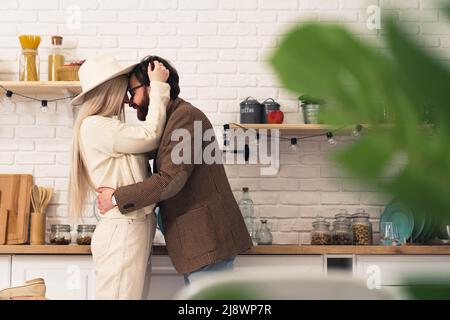 Bell'uomo bearded dai capelli scuri che abbraccia una donna caucasica bionda in piedi in un interno moderno della cucina. Foto di alta qualità Foto Stock
