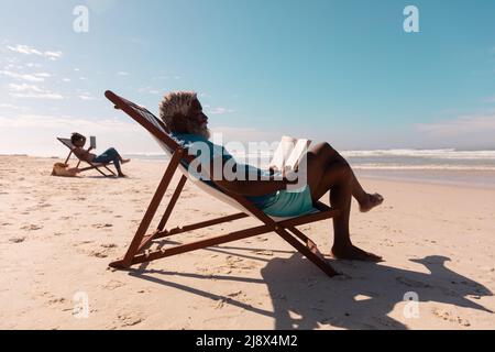 Uomo anziano e donna matura afro-americana che legge libri mentre si rilassano su sdraio in spiaggia Foto Stock