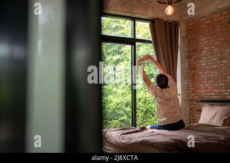 Un uomo pigro felice svegliarsi nel letto alzandosi le mani alla finestra al mattino con fresco sensazione di relax in una camera marrone fatta di mattoni. Foto Stock