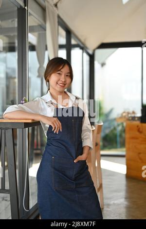 Ritratto, bello e di successo asiatico giovane caffè proprietario in jeans grembiule in piedi nel suo bar. Amichevole barista femminile asiatica in uniforme. Foto Stock